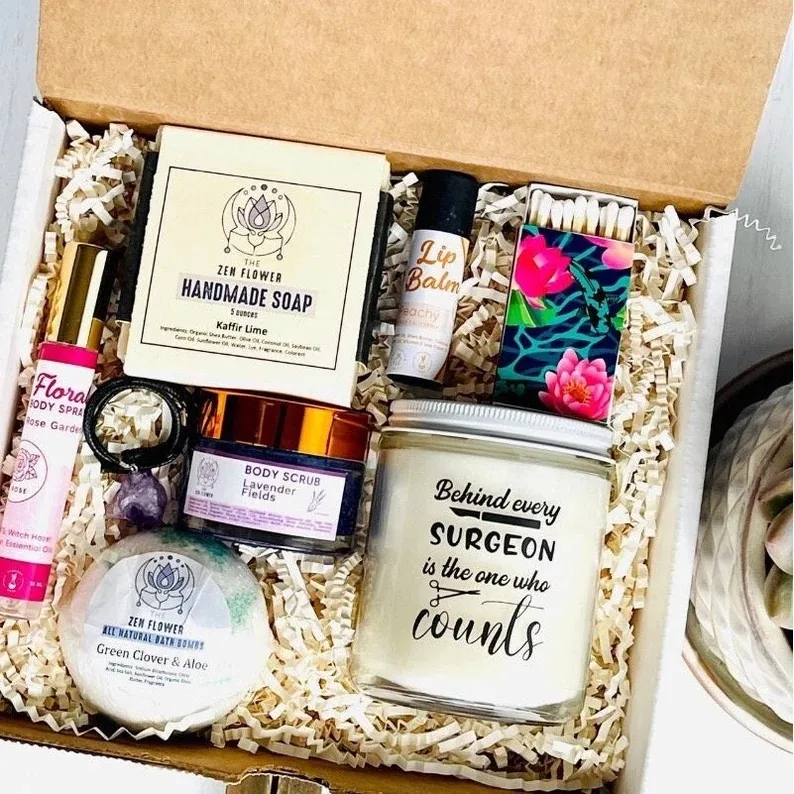 An array of self-care products inside a gift box, including vegan lip balm, sugar scrub, handmade soap, bath bombs, and a candle with the inscription 'Behind every SURGEON is the one who COUNTS', designed for surgical techs' relaxation and care.